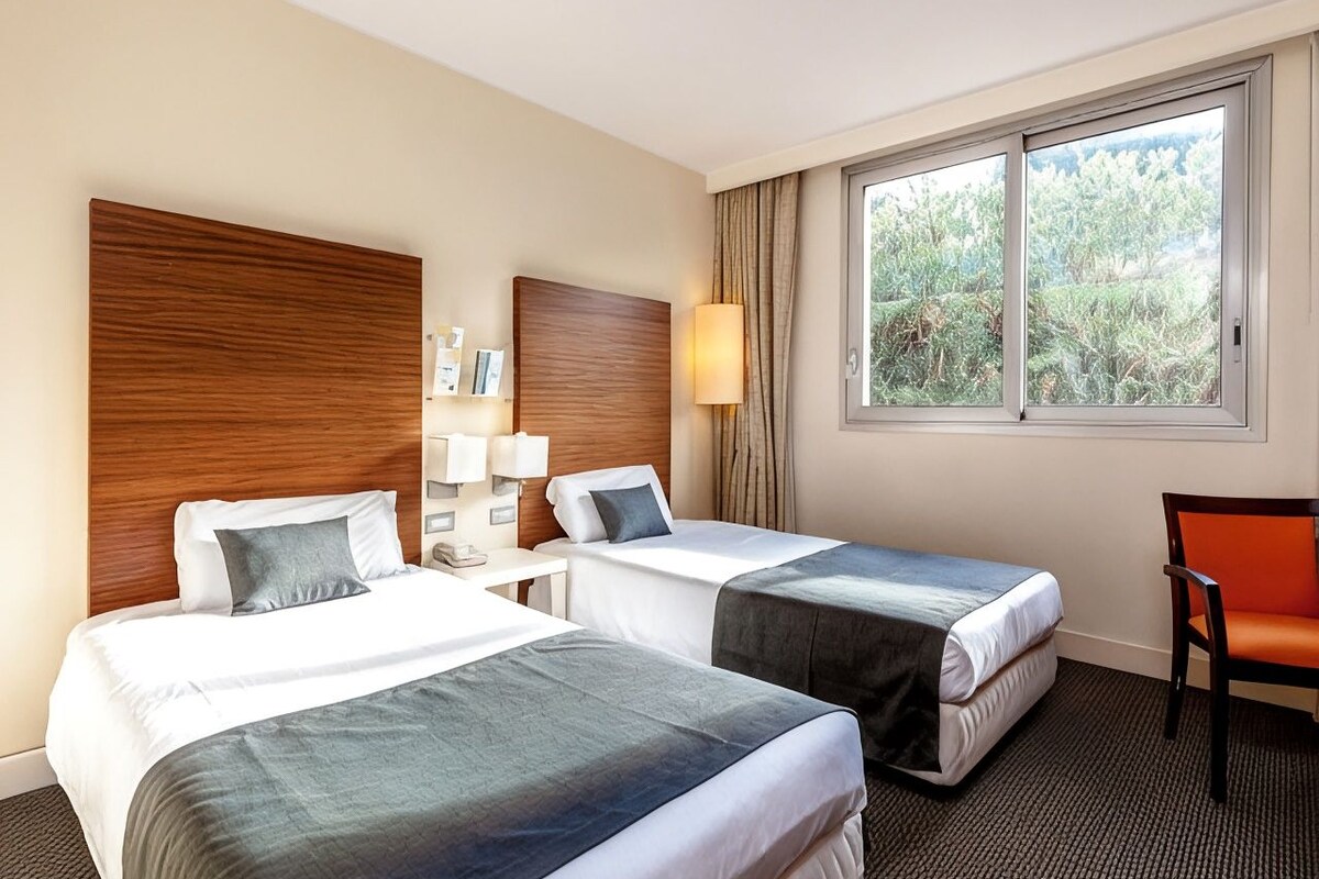 A hotel room with two beds, a desk, a chair, and a window with a view of greenery.