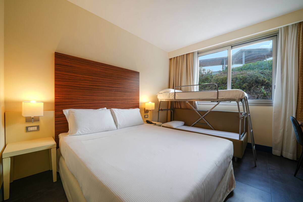 A hotel room with a bunk bed, a bedside table with a lamp, a wooden headboard, and a window with curtains.