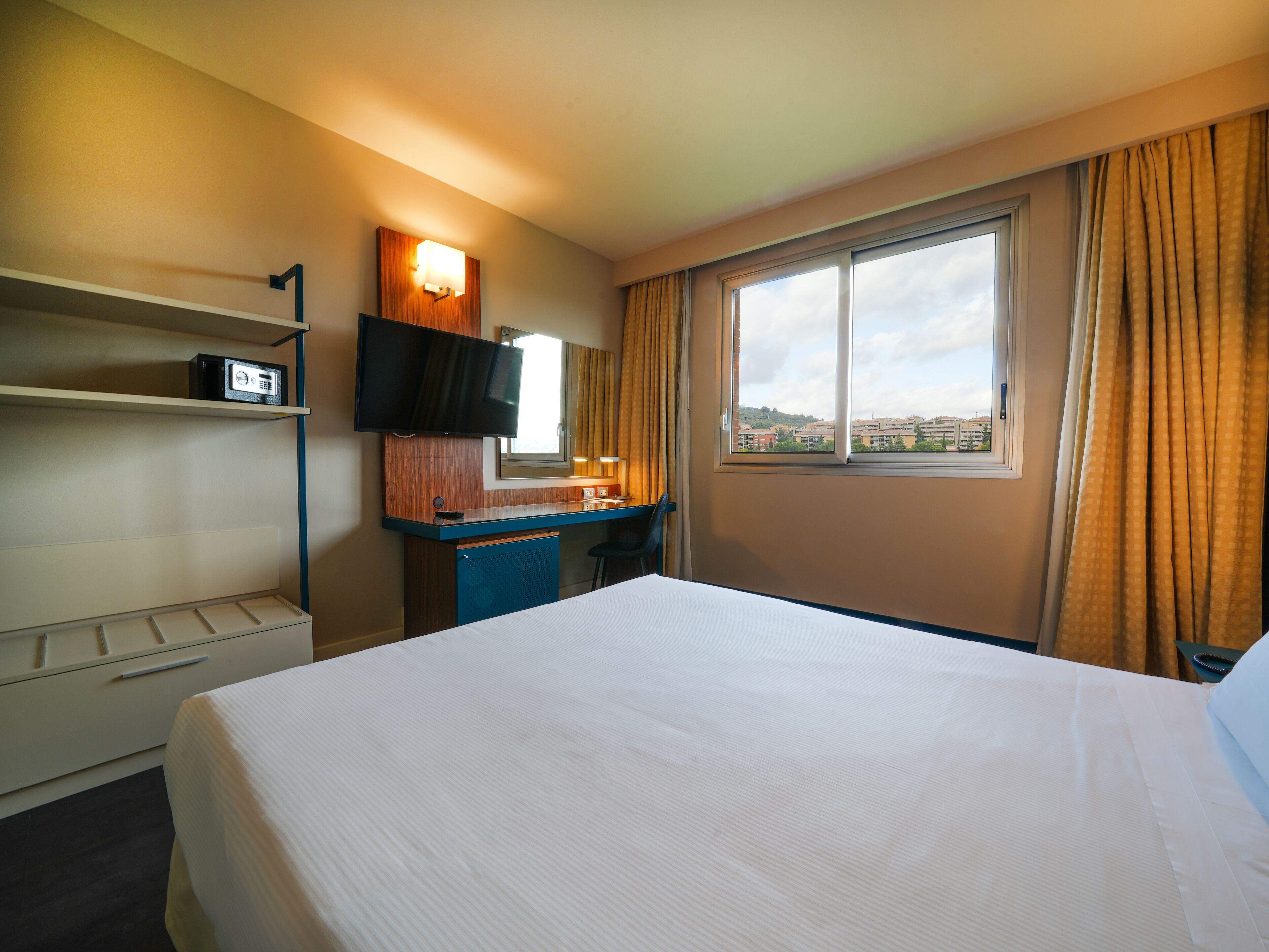 Quarto de hotel com cama, escrivaninha com televisão de tela plana, espelho, janela com cortinas e vista para edifícios.