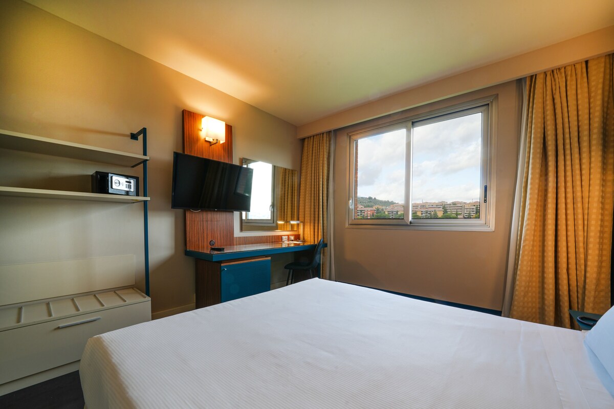 A hotel room with a bed, a desk with a flat-screen TV, a mirror, a window with curtains, and a view of buildings outside.