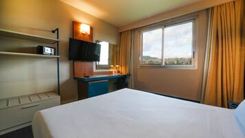 Quarto de hotel com cama, escrivaninha com televisão de tela plana, espelho, janela com cortinas e vista para edifícios.