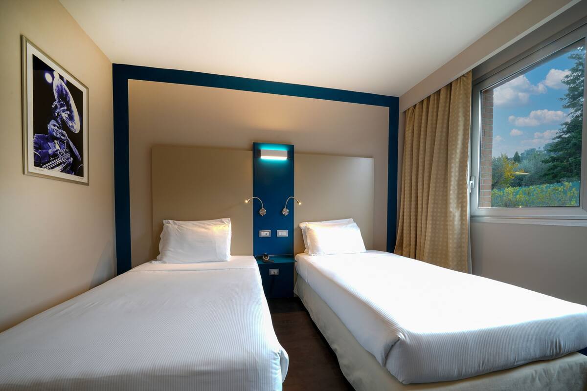 A hotel room with two single beds, a headboard with built-in lighting, a framed picture on the wall, and a window with curtains.