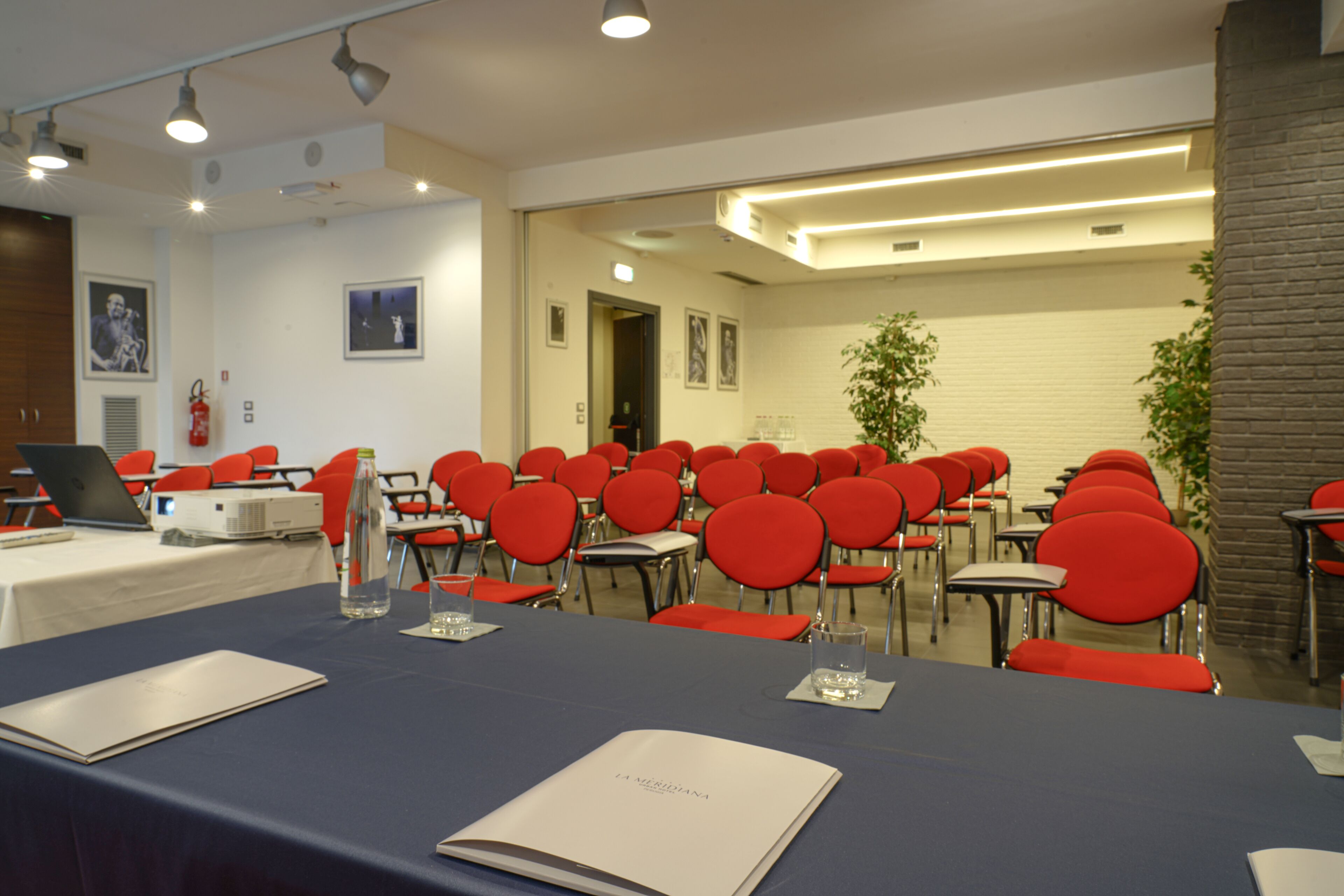 Ein Konferenzraum mit Reihen roter Stühle, einem langen Tisch mit blauer Tischdecke und einem Laptop auf der linken Seite.