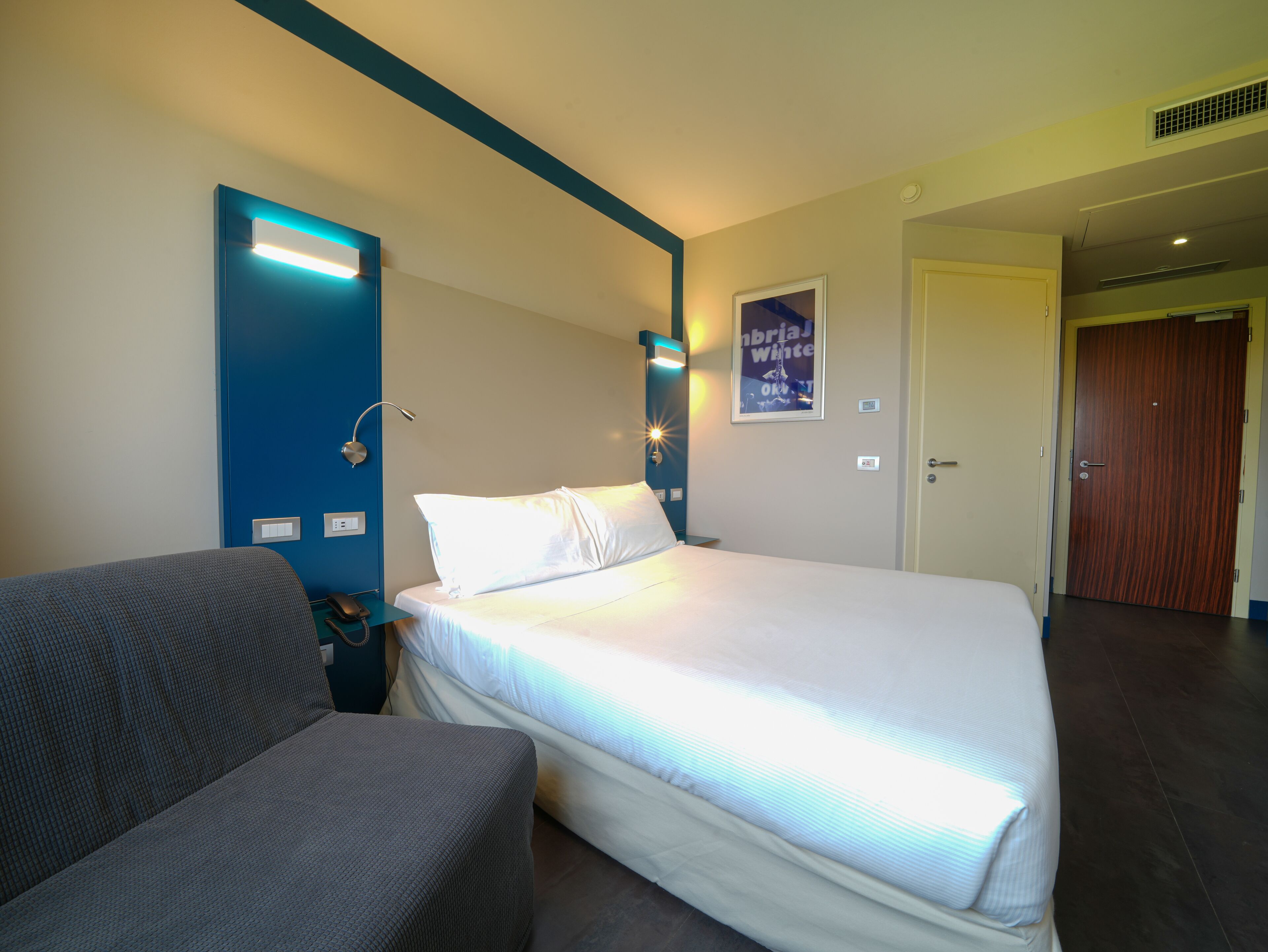 Una habitación de hotel con una cama, una silla, un escritorio con teléfono, una lámpara de pared y un cartel en la pared.
