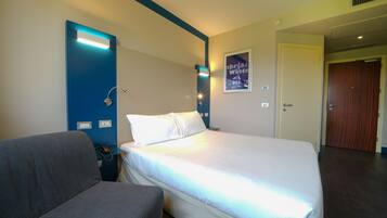 A hotel room with a bed, a chair, a desk with a phone, a wall-mounted lamp, and a poster on the wall.