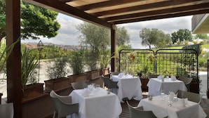 An outdoor dining area with tables set for two, surrounded by greenery and overlooking a scenic view.