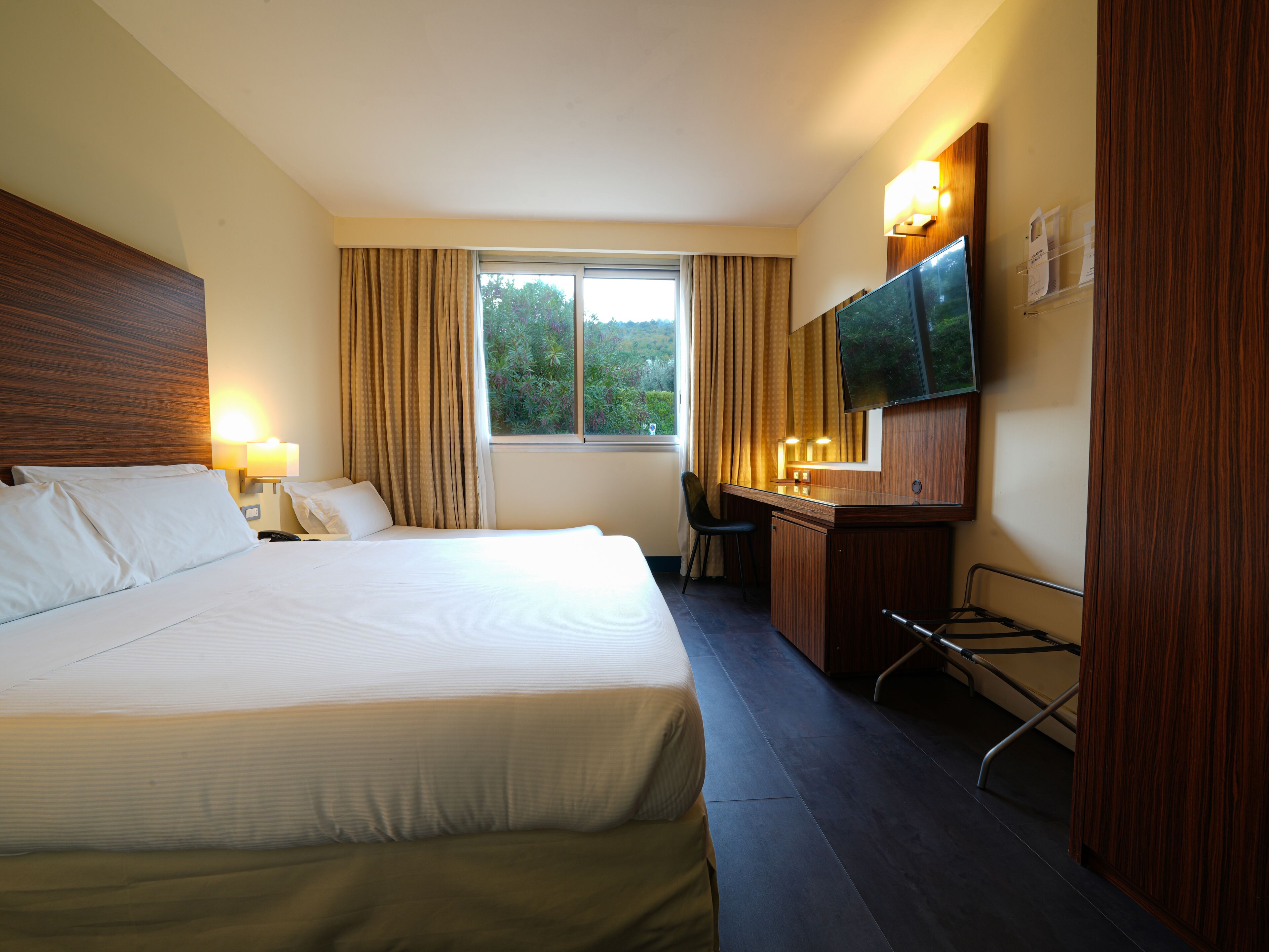 Quarto de hotel com uma cama grande, uma escrivaninha, uma cadeira, uma televisão e uma janela com cortinas.