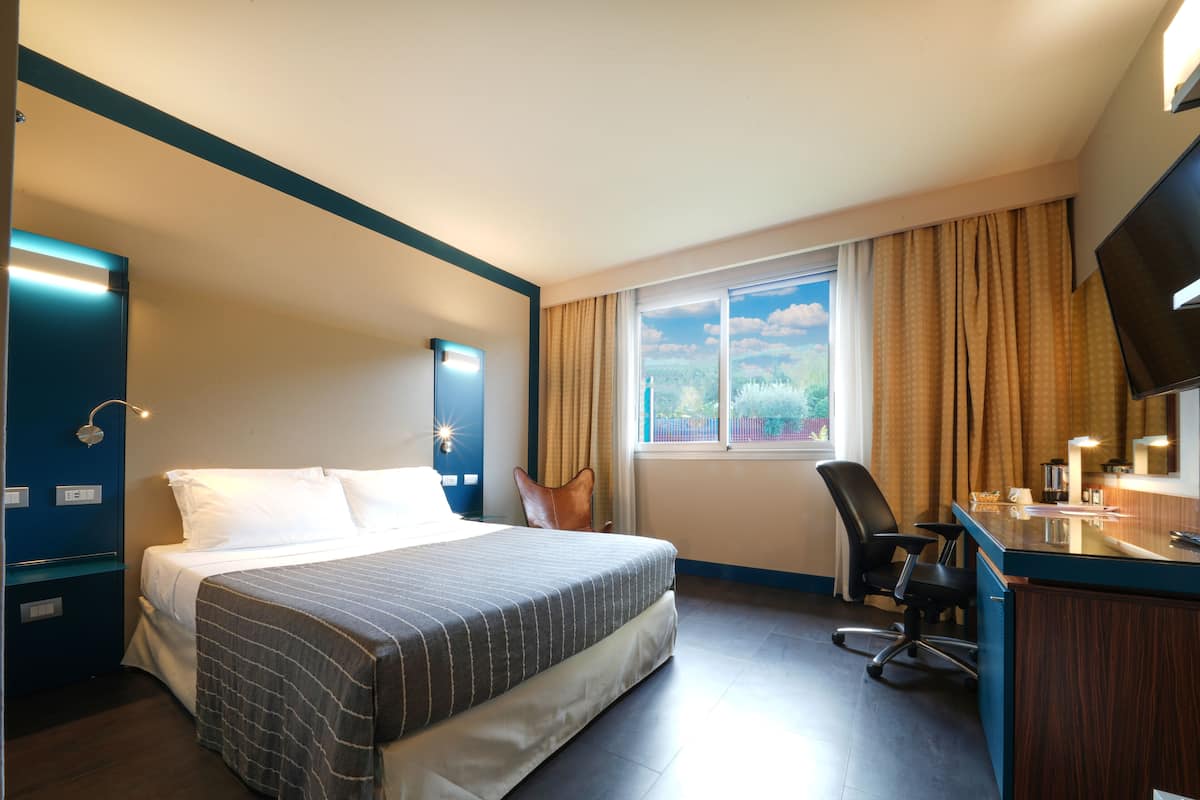 A hotel room with a bed, a desk, a chair, a TV, and a window with curtains.
