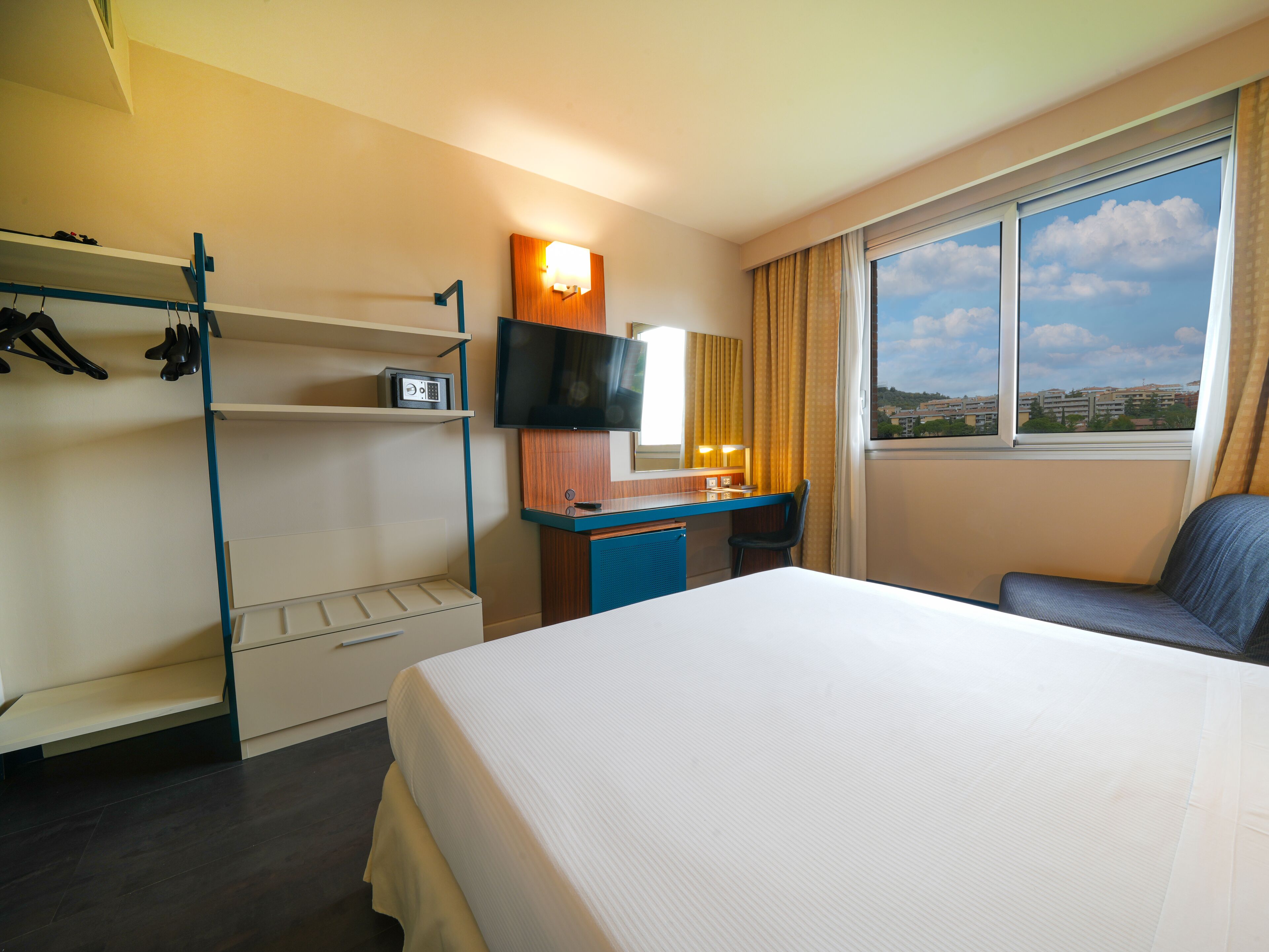 A hotel room with a bed, a desk with a chair, a TV, and a window with a view of buildings.