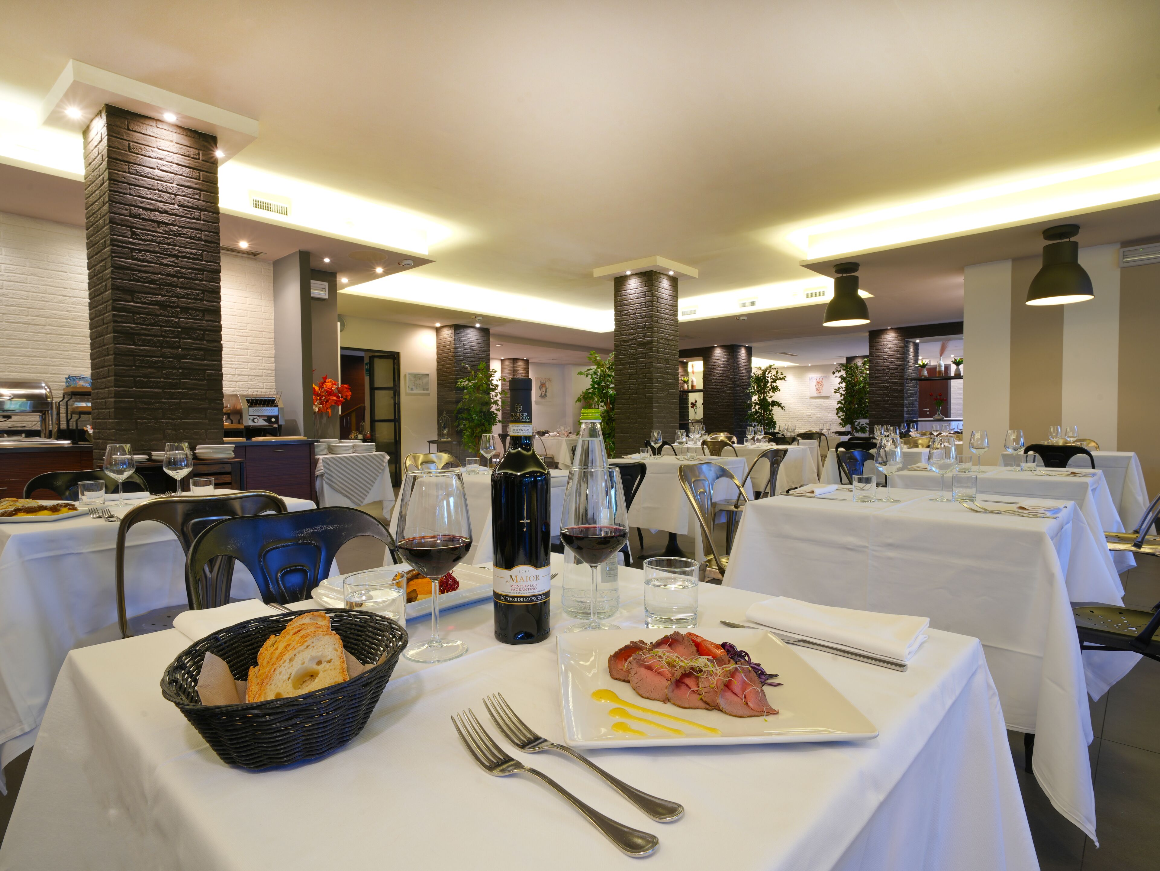 Un restaurante con varias mesas, cada una con mantel blanco, copas de vino y una botella de vino tinto.