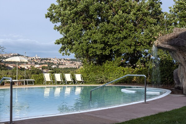 A swimming pool with lounge chairs and a waterfall feature, surrounded by greenery and overlooking a distant town. - La Meridiana Bleisure Hotel (Perugia)