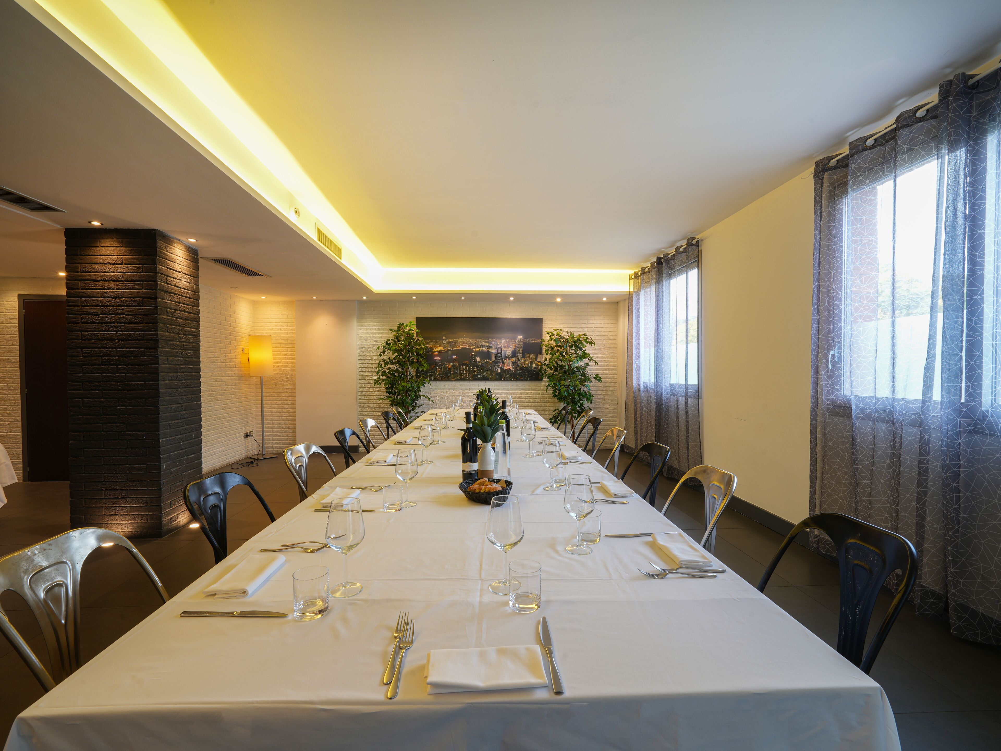 A long dining table set with white tablecloth, chairs, and dinnerware, with a cityscape view through a window.