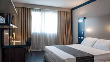 A hotel room with a bed, a desk, a chair, a TV, and a window with curtains.
