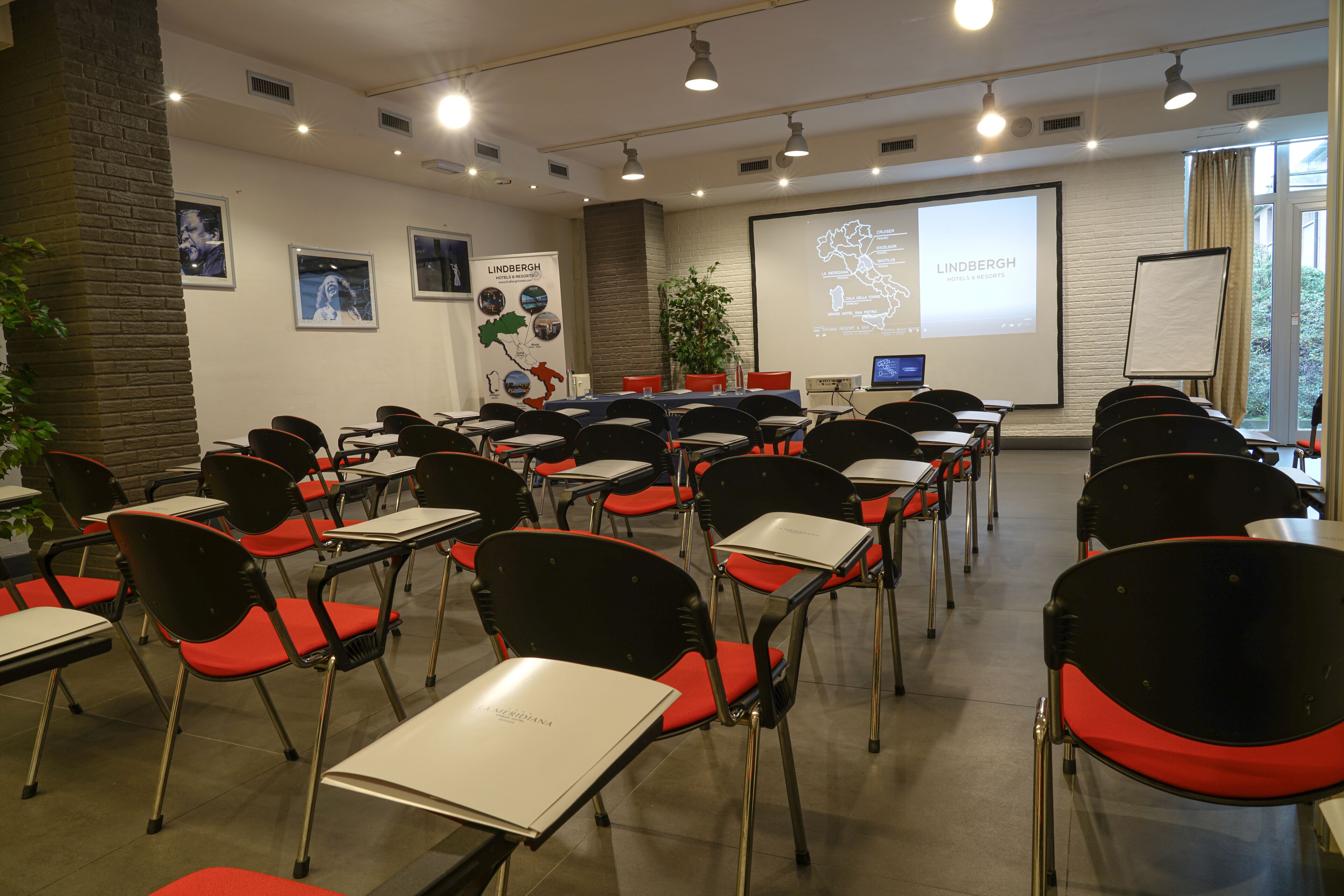 Un salón de conferencias con filas de sillas negras con cojines rojos, mesas blancas, una pantalla de proyector y una presentación sobre Lindbergh.