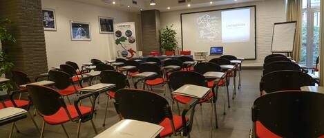 Un salón de conferencias con filas de sillas negras con cojines rojos, mesas blancas, una pantalla de proyector y una presentación sobre Lindbergh.