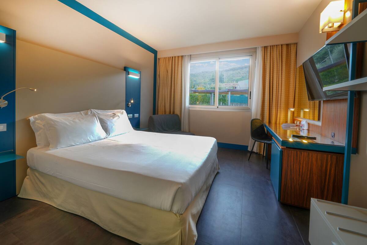 A hotel room with a bed, a desk, a chair, a TV, and a window with a view.
