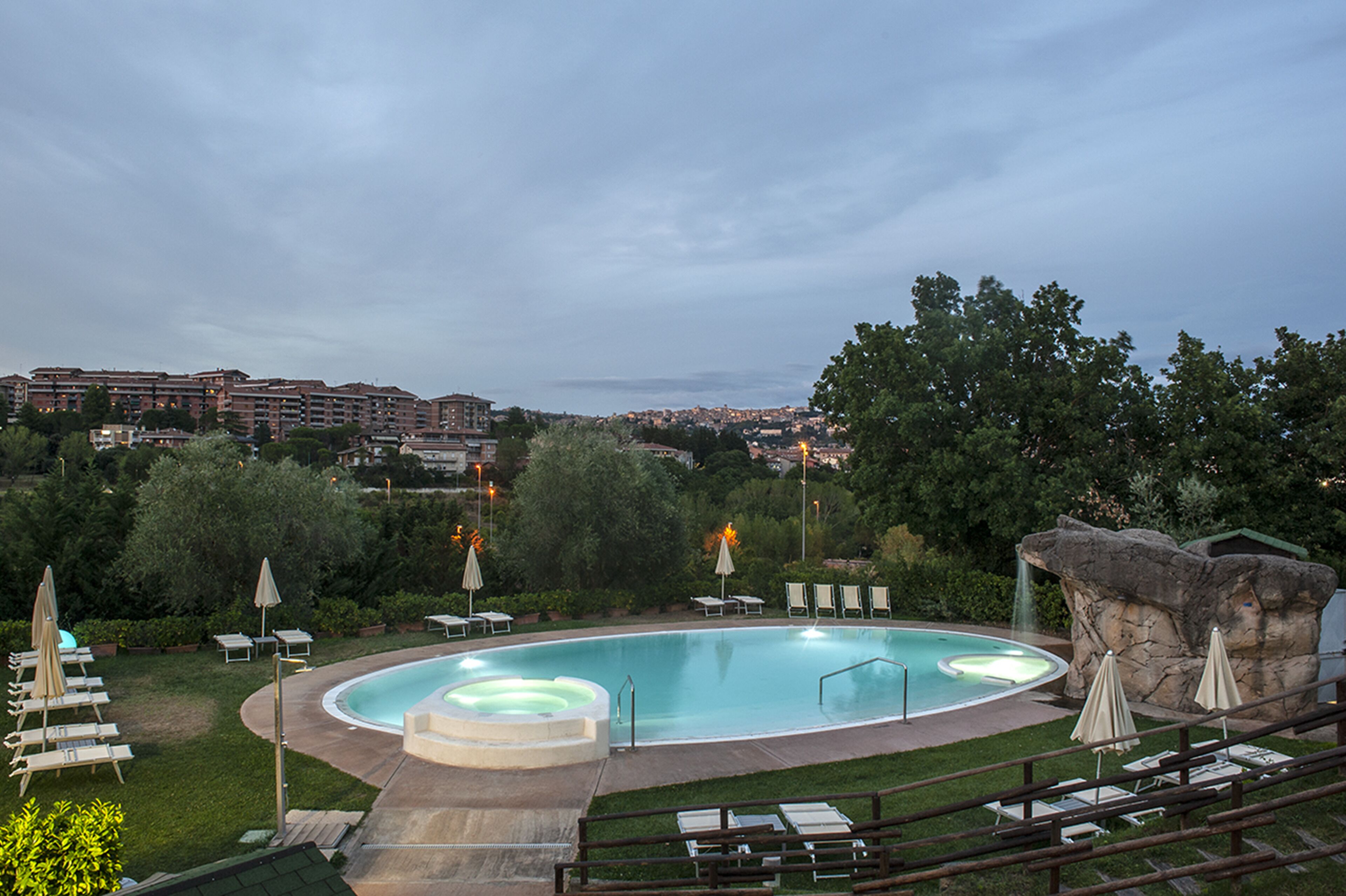 A swimming pool with lounge chairs and umbrellas, surrounded by greenery and a view of a cityscape in the background.
