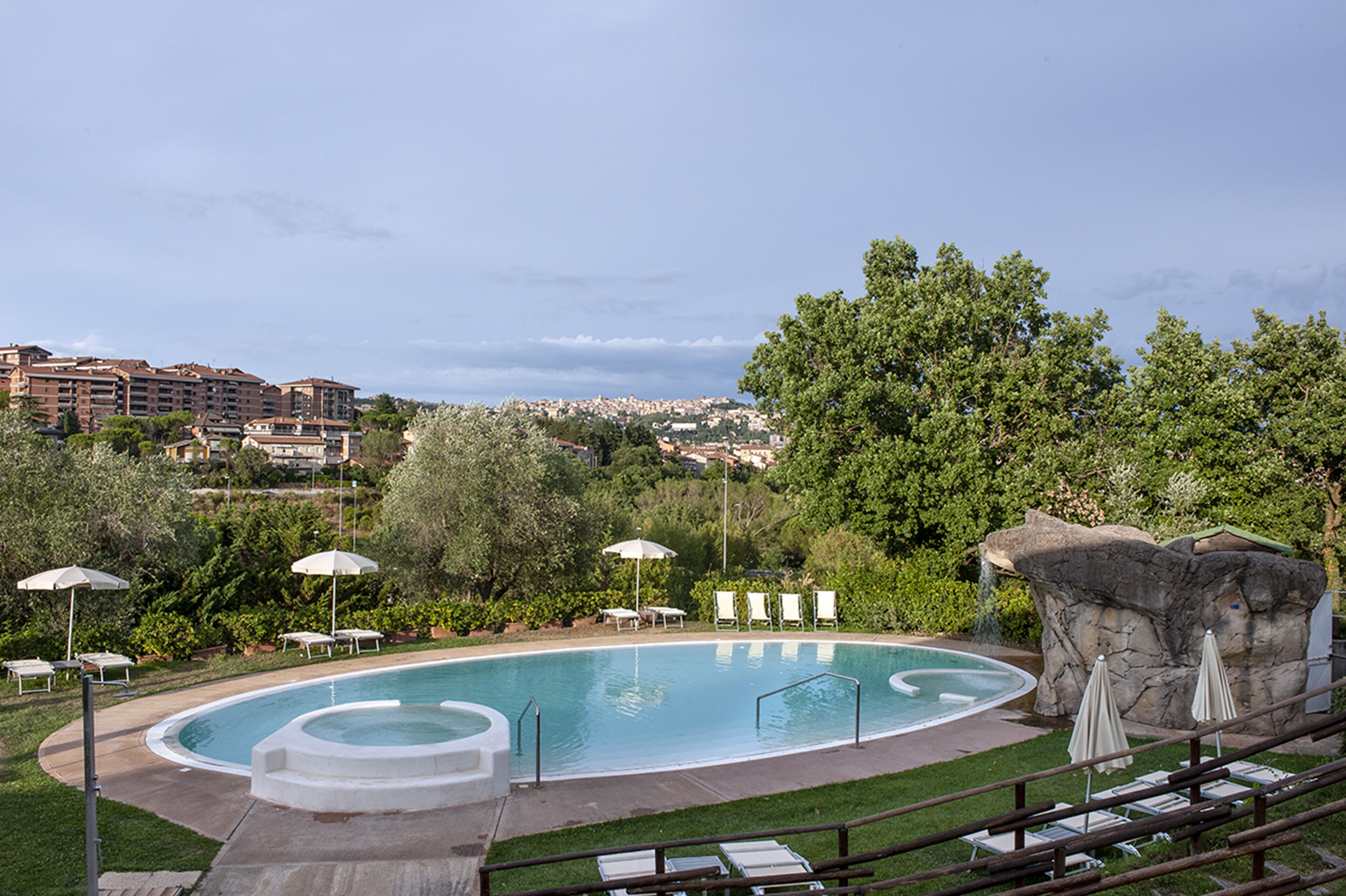A swimming pool with lounge chairs and umbrellas, surrounded by greenery and a view of a cityscape in the background.