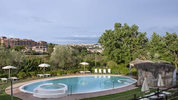 Una piscina con sillones de descanso y sombrillas, rodeada de vegetación y vistas a un paisaje urbano al fondo.