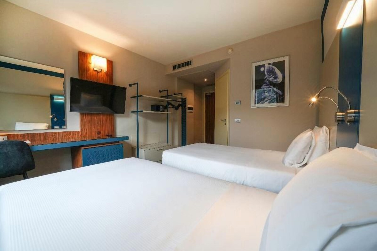 A hotel room with two beds, a desk with a chair, a flat-screen TV, a mirror, and a wall-mounted light fixture.