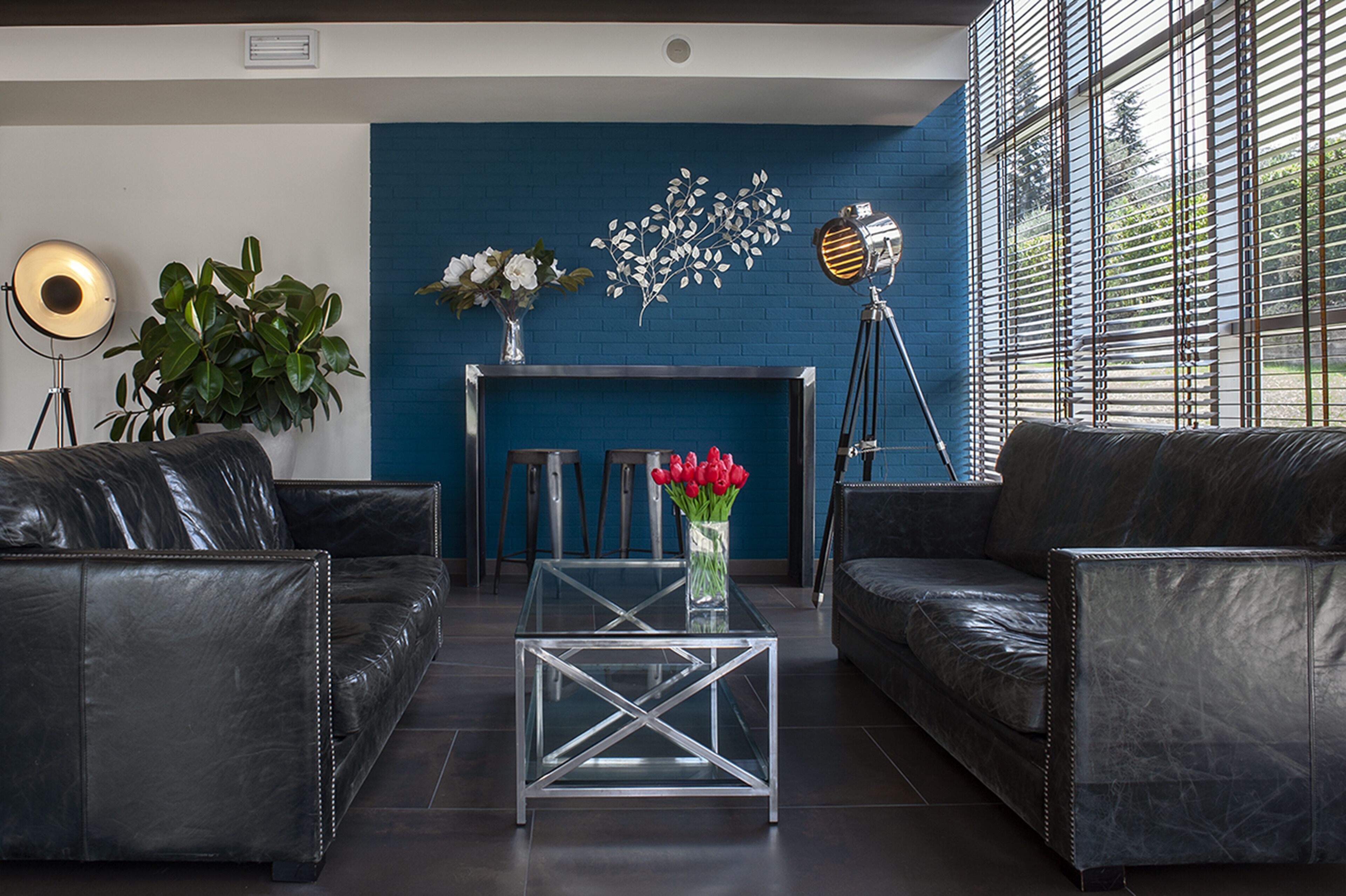 A modern living room with two black leather sofas, a glass coffee table, a blue brick wall, a white fireplace, and a large window with blinds.