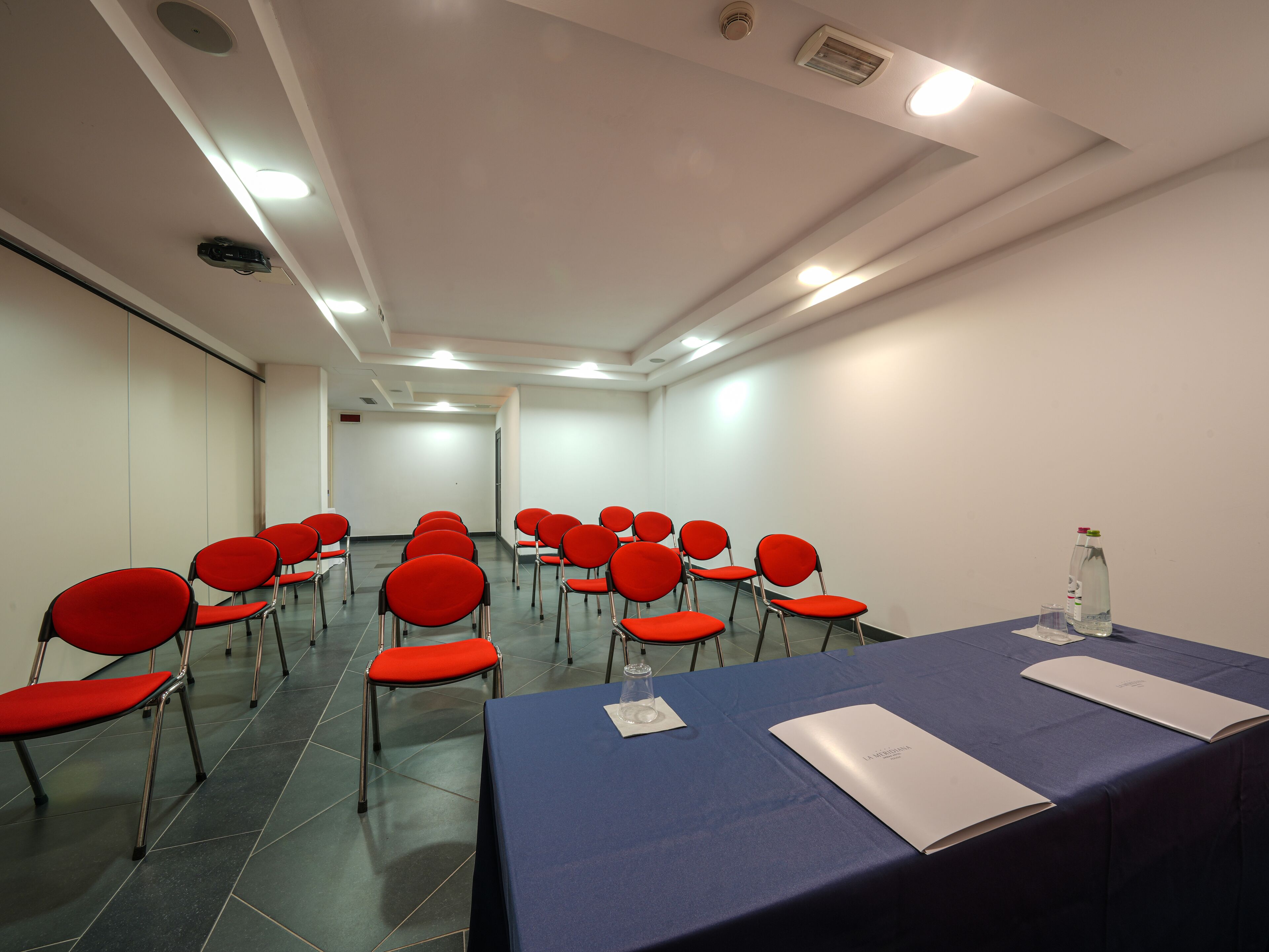 A conference room with rows of red chairs, a blue table with documents, and a water bottle.