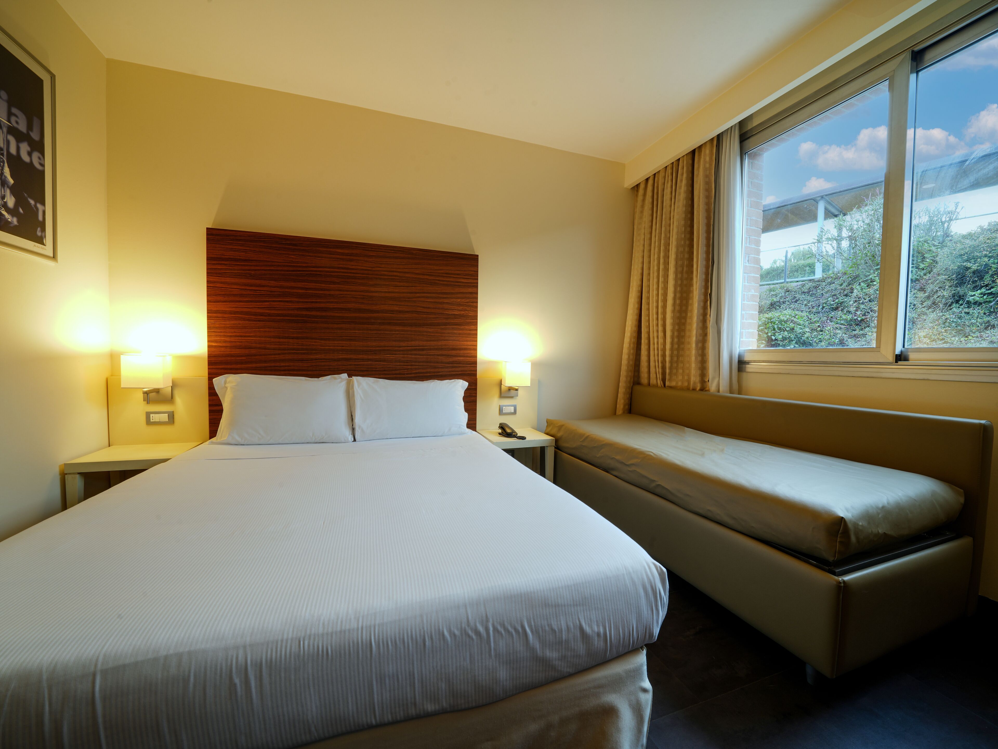 A hotel room with a large bed, bedside tables, a bench, and a window with a view of greenery.