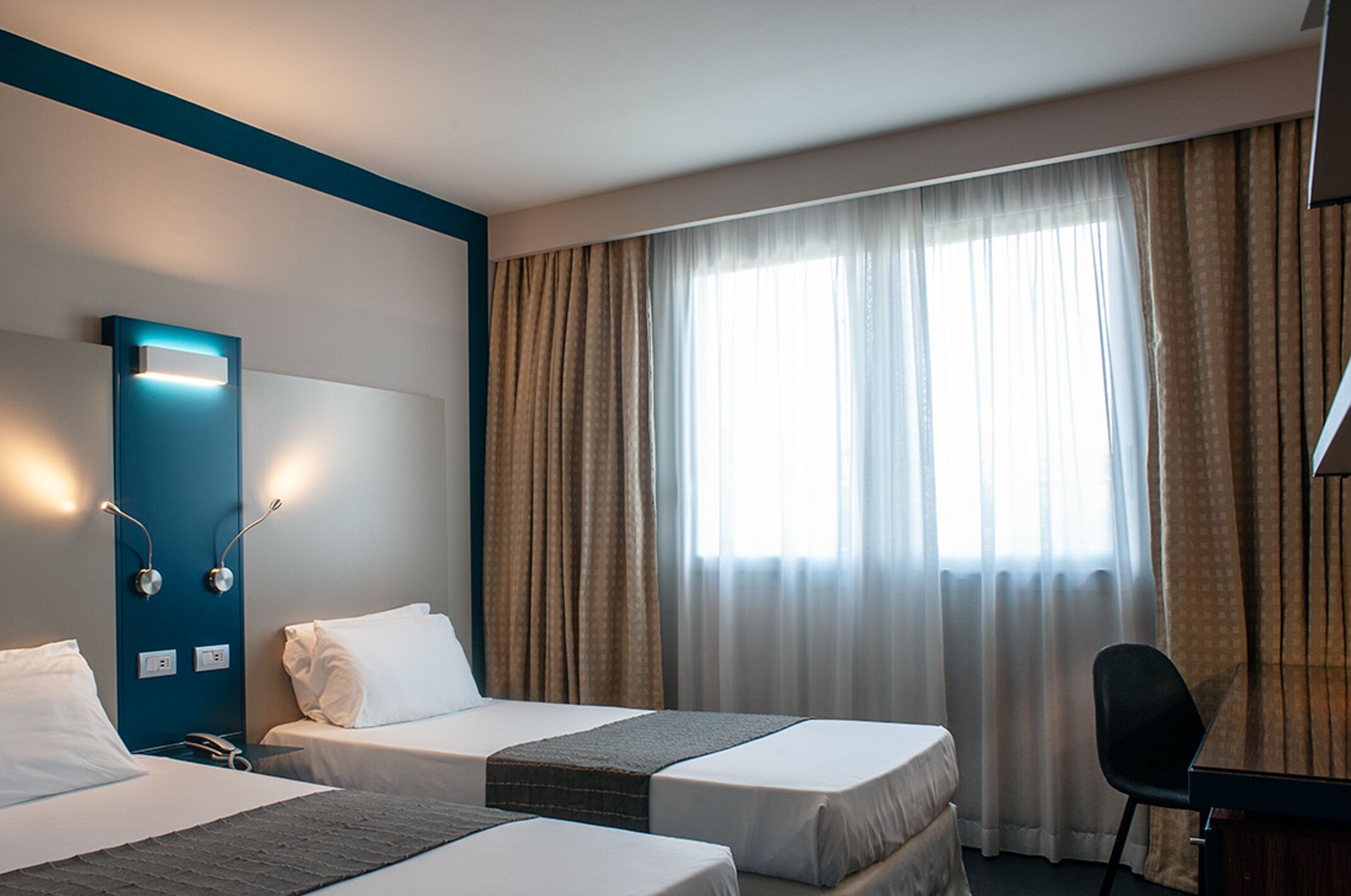 A hotel room with two beds, a desk, and a large window with curtains.