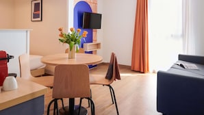 Une chambre d’hôtel moderne avec une table à manger ronde, un canapé, une télévision et une grande fenêtre avec des rideaux.