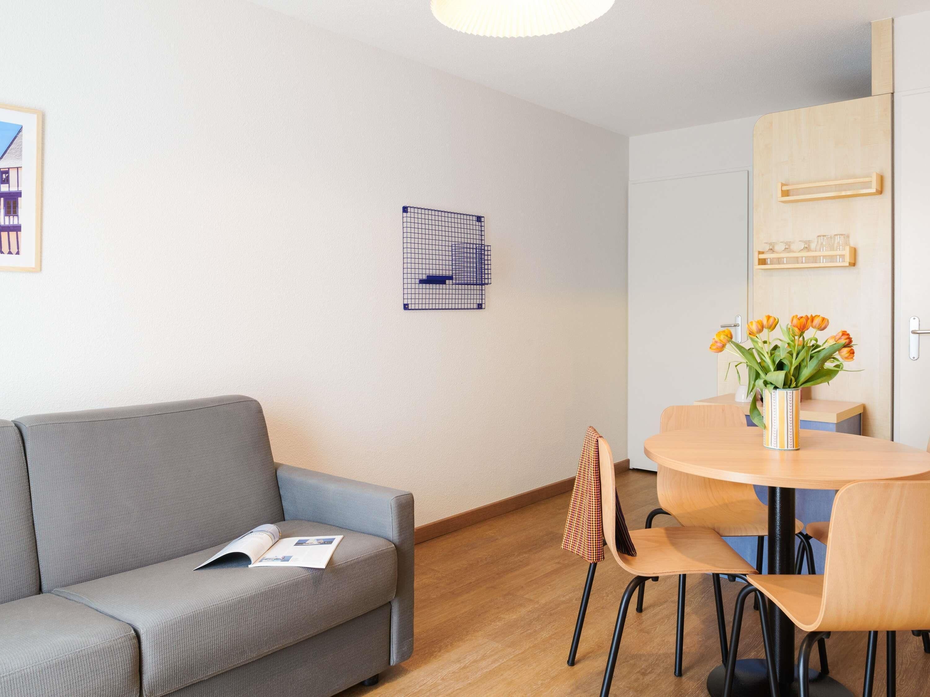 Una habitación moderna con un sofá gris, una mesa de comedor de madera con sillas, un jarrón de flores y un cuadro colgado en la pared.