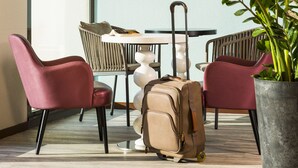 Une chambre d’hôtel moderne avec un grand téléviseur à écran plat fixé au mur, une table à manger avec des chaises, une plante en pot et une valise posée sur le sol.