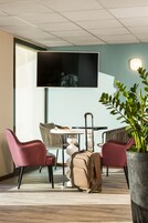 Une chambre d’hôtel moderne avec un grand téléviseur à écran plat fixé au mur, une table à manger avec des chaises, une plante en pot et une valise posée sur le sol.