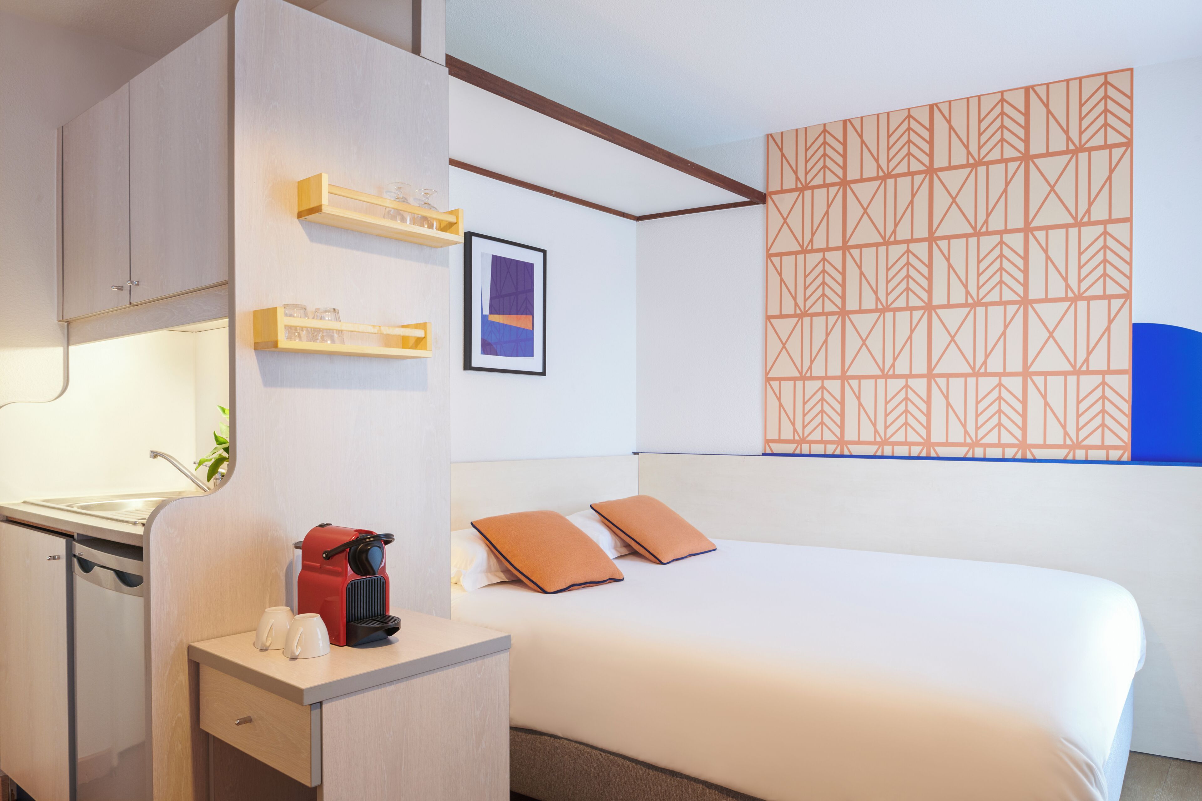 A hotel room with a bed, a desk with a coffee maker, a small cabinet, and a wall with a geometric pattern.