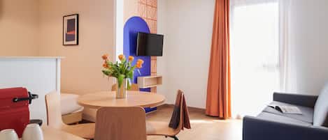 A modern hotel room with a dining area, a flat-screen TV, a sofa, and a large window with curtains.