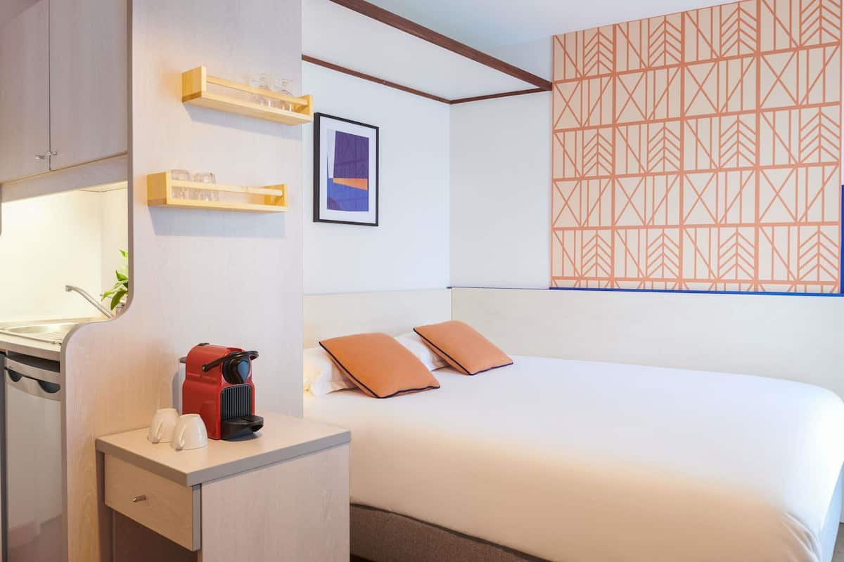 A hotel room with a bed, a desk with a coffee maker, a shelf with glasses, and a geometric-patterned wall.