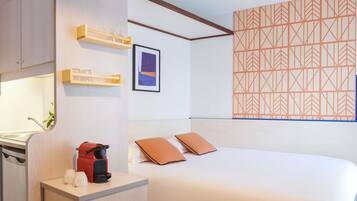 A hotel room with a bed, a desk with a coffee maker, a shelf with glasses, and a geometric-patterned wall.
