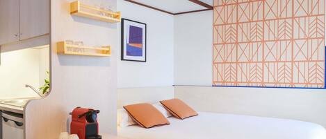 A hotel room with a bed, a desk with a coffee maker, a shelf with glasses, and a geometric-patterned wall.