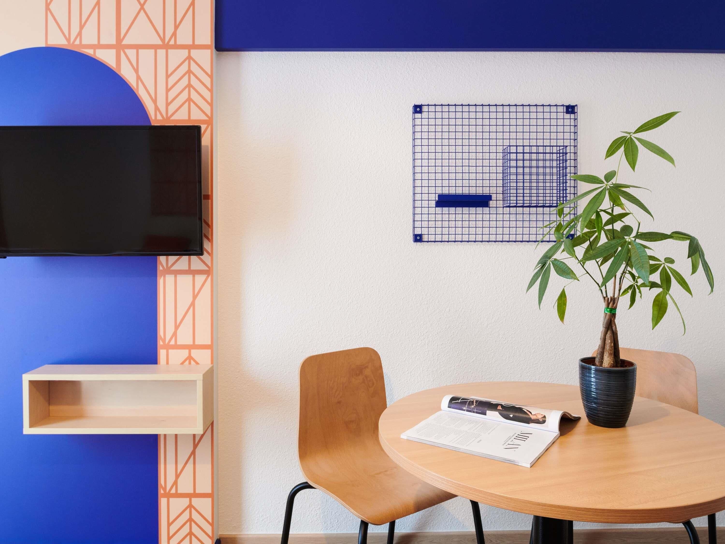 A modern room with a round wooden table, a potted plant, a flat-screen TV mounted on the wall, and a blue geometric-patterned shelf.