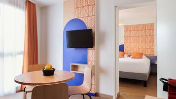 Habitación de hotel con cama, televisión montada en la pared, mesa de comedor con sillas y una cesta de frutas.