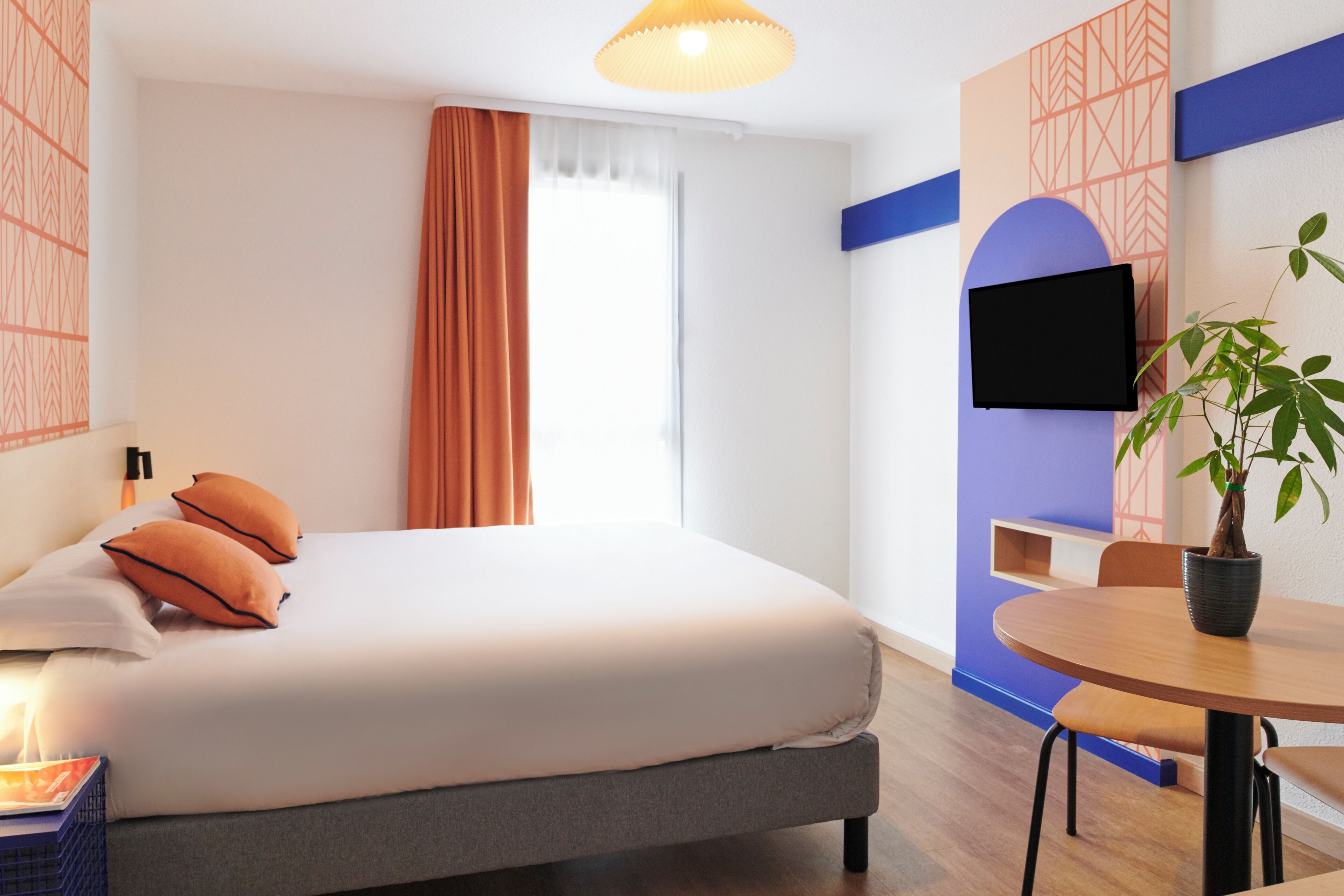 A modern hotel room with a bed, a TV mounted on the wall, a dining table, and a potted plant.