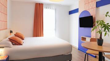 A modern hotel room with a bed, a TV mounted on the wall, a dining table, and a potted plant.