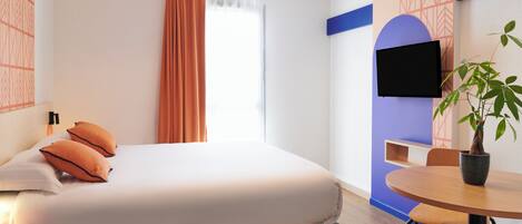 A modern hotel room with a bed, a TV mounted on the wall, a dining table, and a potted plant.