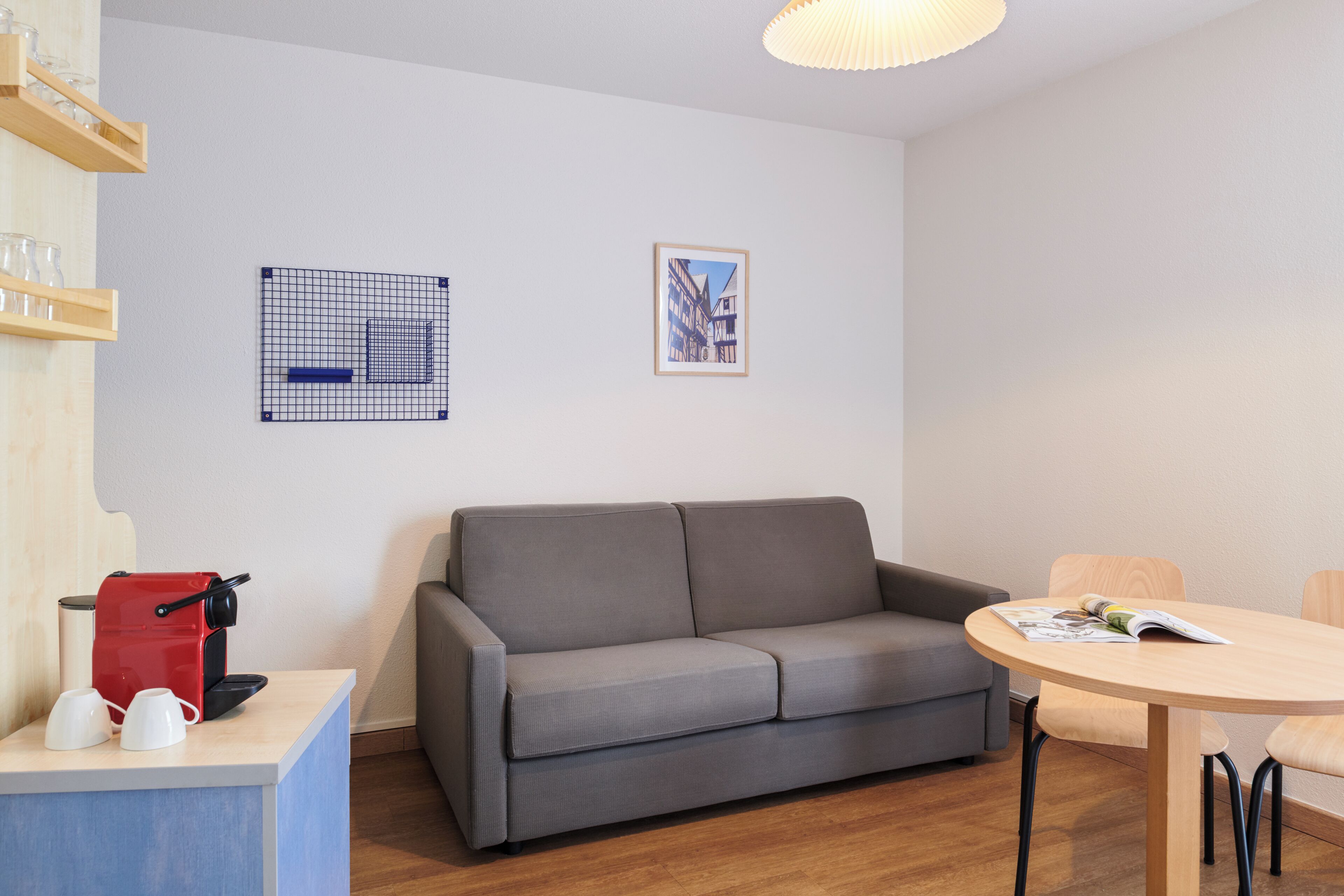 Un salon moderne avec un canapé gris, une table basse en bois et une petite table à manger. Deux tableaux encadrés ornent le mur, ainsi qu’une machine à café et un ensemble de verres posés sur le comptoir.
