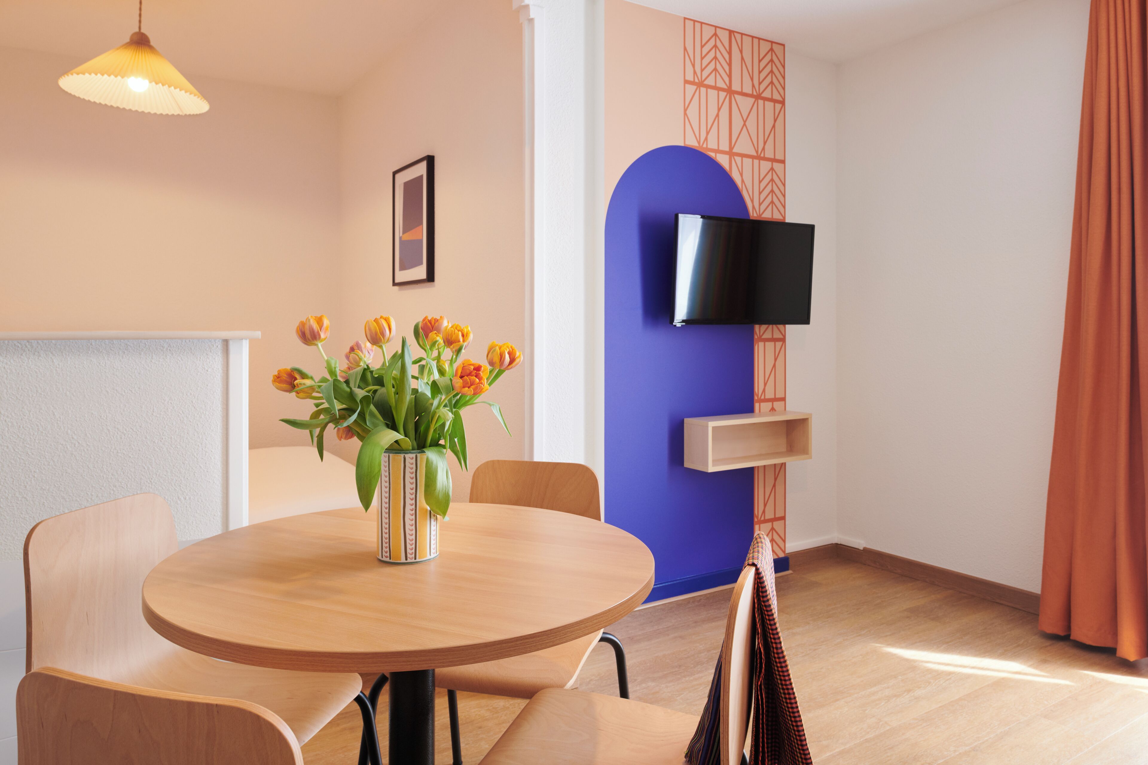 A room with a round wooden table, chairs, a vase with flowers, a television on the wall, and a patterned wall design.