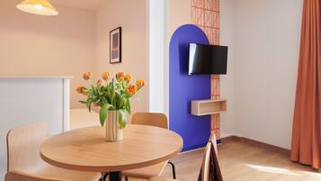 A room with a round wooden table, chairs, a vase with flowers, a television on the wall, and a patterned wall design.