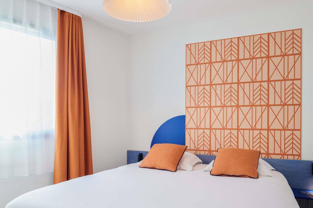 A hotel room with a bed, curtains, a patterned wall, and a ceiling light.