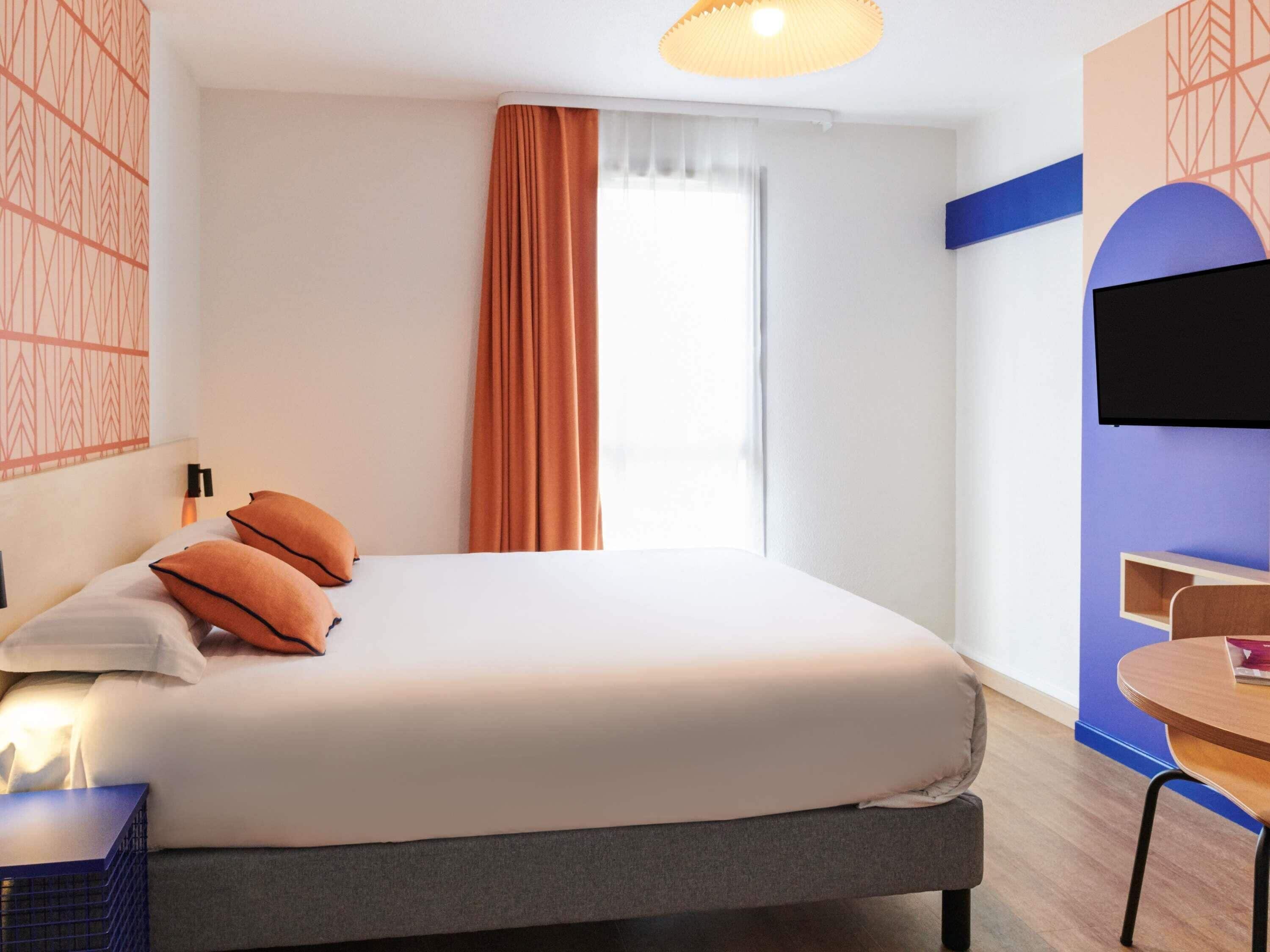 A hotel room with a bed, a desk, a chair, a TV, and a window with curtains.