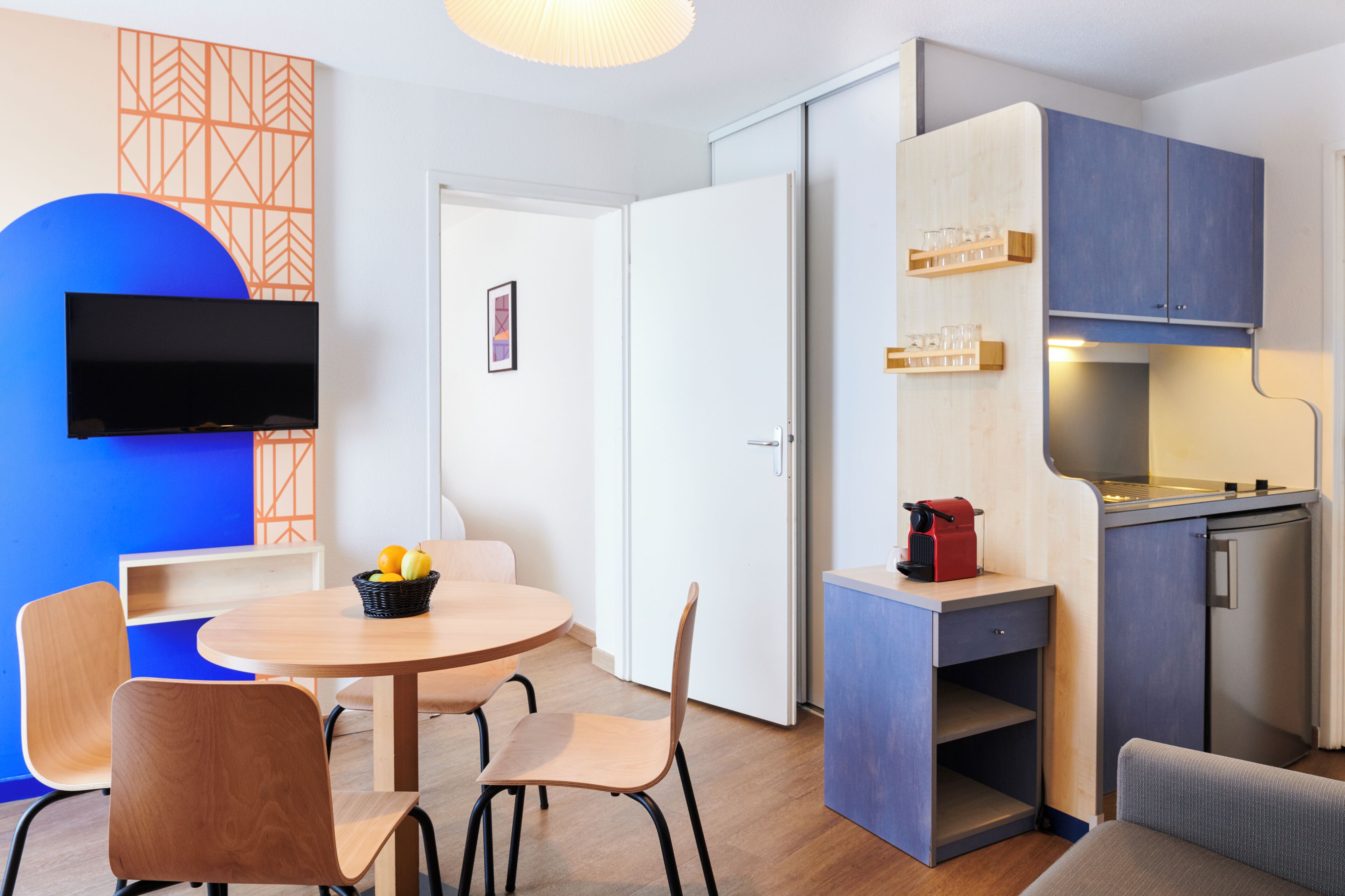 Un espacio habitable compacto con una mesa redonda de comedor, sillas, un televisor montado en la pared, una kitchenette y una puerta que da a otra habitación.