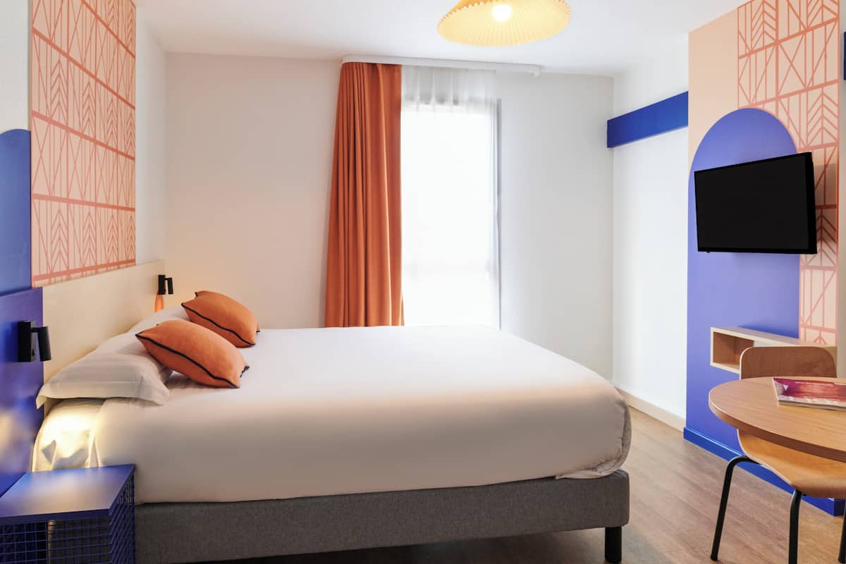 A hotel room with a bed, a desk, a chair, a TV, and a window with curtains.