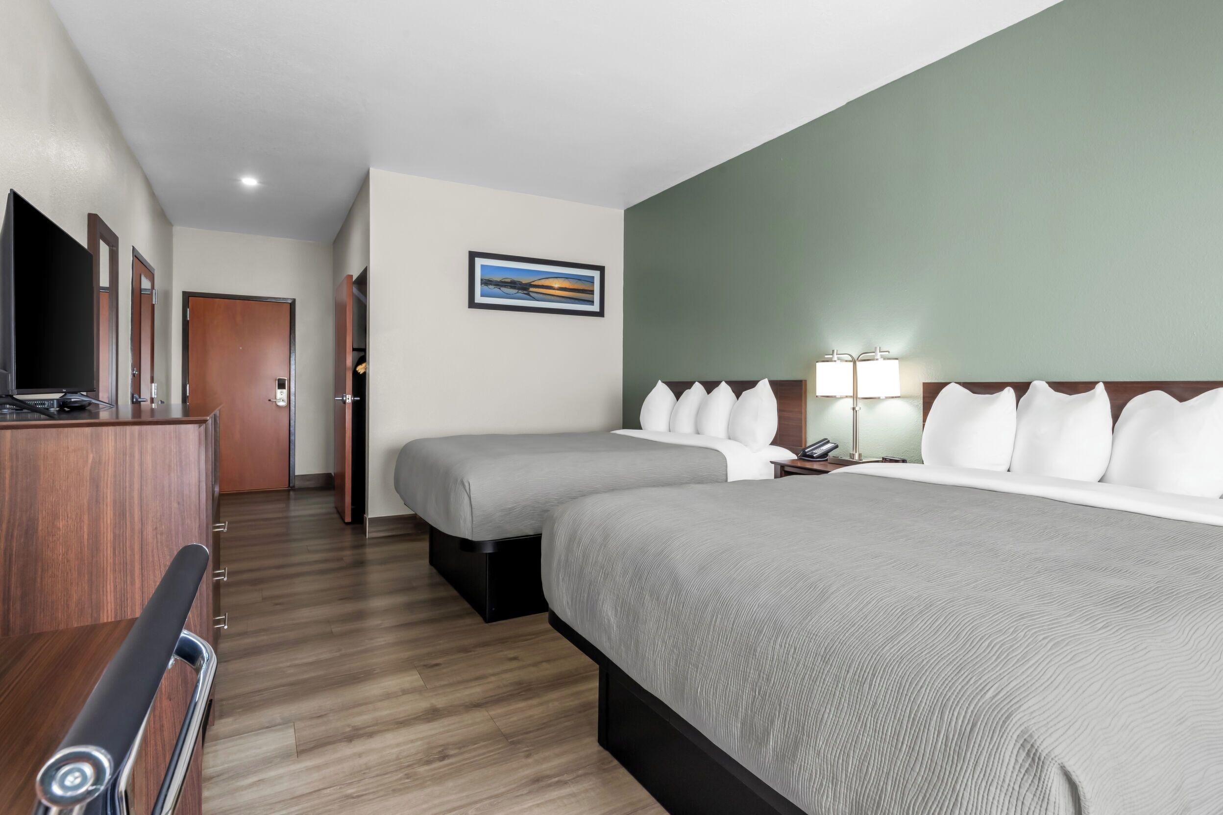 A hotel room with two beds, a desk with a TV, a painting on the wall, and a lamp on the nightstand.